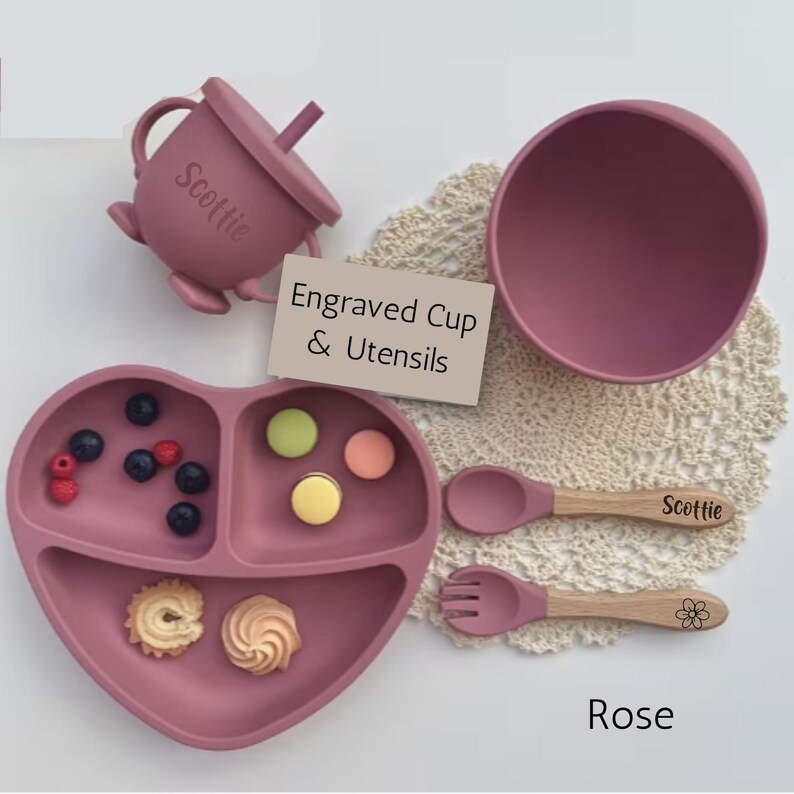 May include: A rose-colored silicone baby feeding set, including a heart-shaped divided plate, bowl, cup with lid, spoon, and fork. The spoon and cup are engraved with a name. The plate contains berries and cookies.