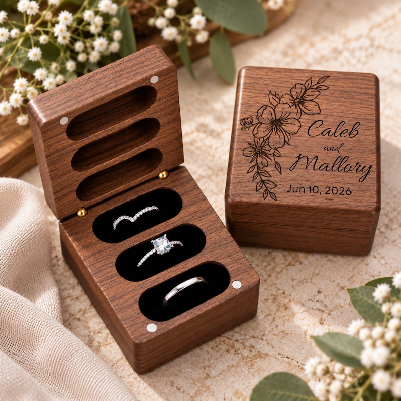 May include: A dark wood ring box with a hinged lid featuring a floral engraving and the text Caleb and Mallory Jun 10, 2026. The open box interior has three black velvet-lined slots containing a chevron ring, a square-cut ring, and a plain band.