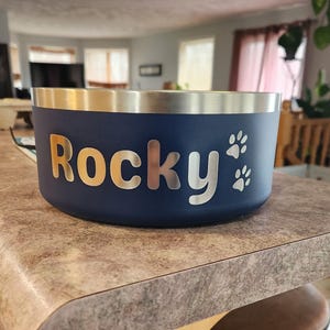 May include: A dark blue pet bowl with a metallic silver rim. The side of the bowl features the name Rocky in a bold, silver-toned font, accompanied by two small silver paw print icons positioned to the right of the name on the matte surface.