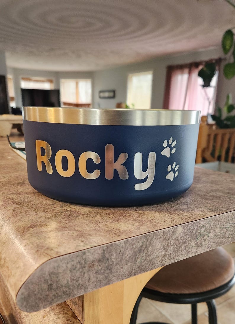 May include: A dark blue pet bowl with a metallic silver rim. The side of the bowl features the name Rocky in a bold, silver-toned font, accompanied by two small silver paw print icons positioned to the right of the name on the matte surface.
