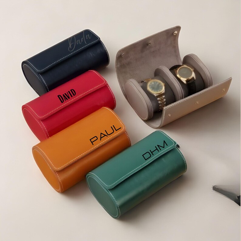 May include: A collection of cylindrical watch travel cases in navy, red, orange, and green, each featuring white stitching and personalised names or initials. One beige open case displays two wristwatches on individual padded cushions inside.