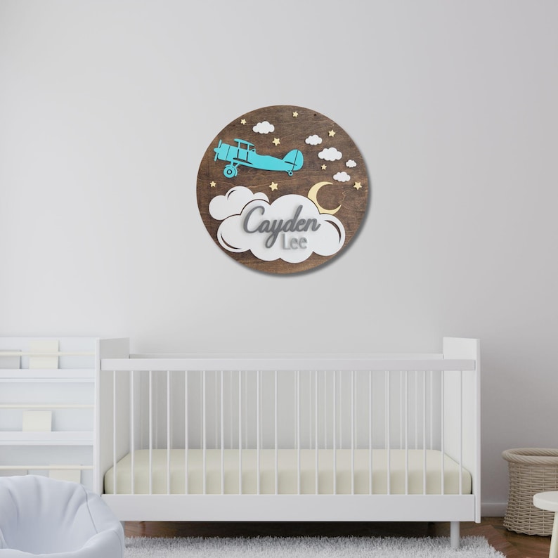 May include: A round wooden wall decor piece featuring a blue airplane, clouds, stars, and a crescent moon. The name "Cayden Lee" is displayed in a cloud-shaped design. The background is a dark brown wood.
