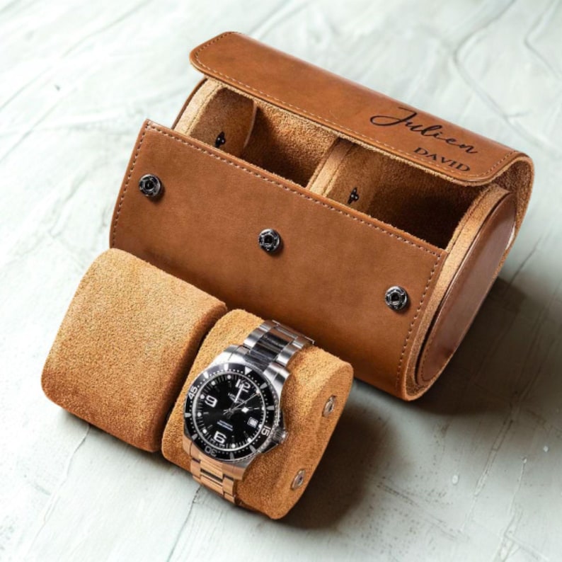 May include: A tan leather watch roll case with a snap button closure and the engraved name Julien David on the flap. The case is open, revealing two soft tan suede watch pillows, one of which holds a silver-toned wristwatch with a black dial.