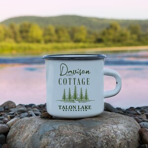Peut inclure: Mug en émail blanc avec un bord et une anse noirs. Le mug porte l'inscription "Davison Family Cottage" au-dessus d'une ligne de pins verts et les mots "Talon Lake, Rutherglen". Le mug est posé sur un rocher avec un arrière-plan flou de lac et de montagne.