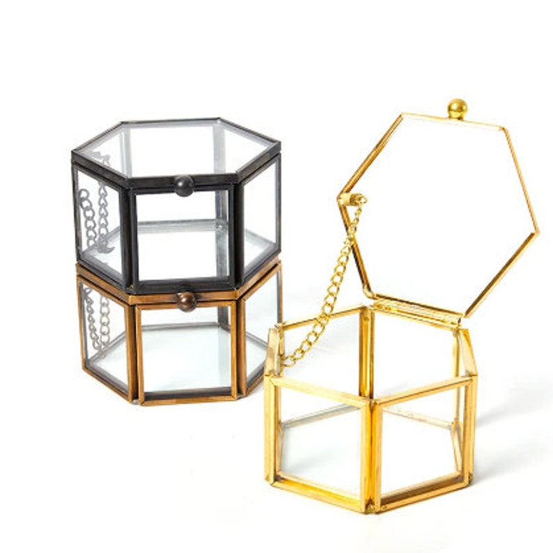 K&ouml;nnte beinhalten: Drei sechseckige Glas-Pr&auml;sentationsboxen mit Metallrahmen. Eine Box ist schwarz, eine bronzefarben und eine goldfarben. Jede Box verf&uuml;gt &uuml;ber einen Klappdeckel, der mit einer kleinen Metallkette befestigt ist, um ihn offen zu halten.
