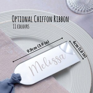 May include: A clear acrylic place card with the name "Melissa" in a script font. The card is 9.9cm long and 4cm wide. The text "Optional Chiffon Ribbon 11 Colours" is above the card. The card is on a white plate with a blue napkin.