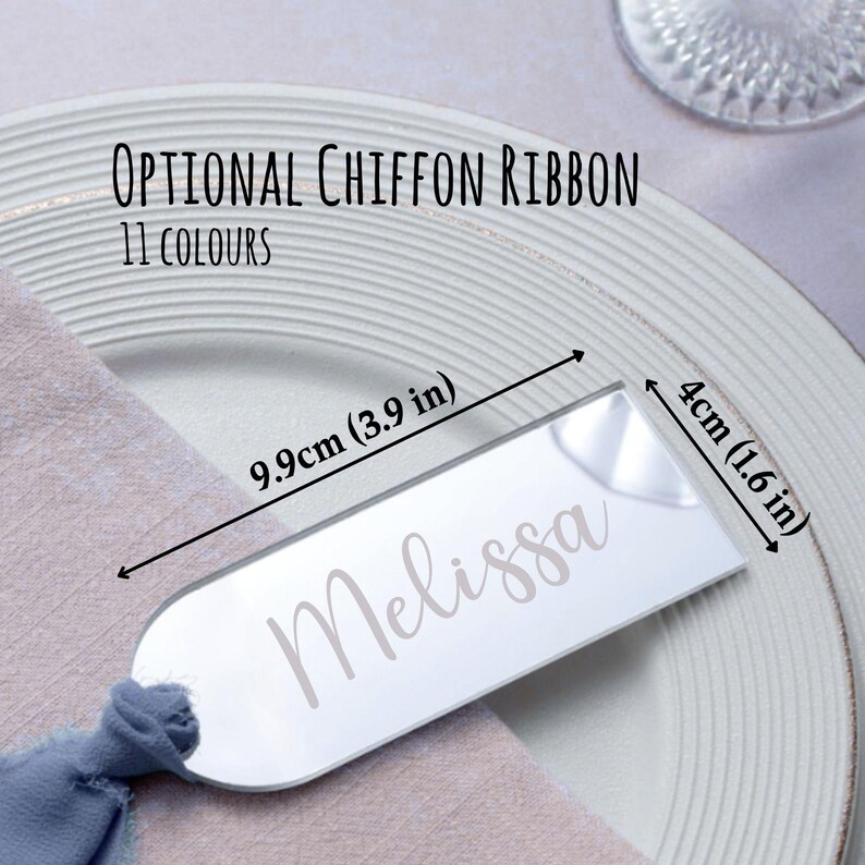 May include: A clear acrylic place card with the name "Melissa" in a script font. The card is 9.9cm long and 4cm wide. The text "Optional Chiffon Ribbon 11 Colours" is above the card. The card is on a white plate with a blue napkin.