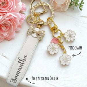 Op de afbeelding: Een witte leren sleutelhanger met de naam "Samantha" in script. De sleutelhanger heeft een gouden strik met kristallen accenten en gouden hardware. Er zijn drie witte en roze bloemvormige bedels aan bevestigd.