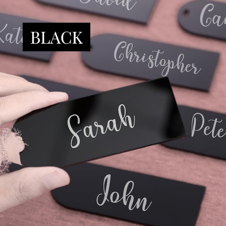 May include: Black name tags with white script lettering. The tags are in various shapes, including rectangular and rounded. One tag is being held by a hand. The word "BLACK" is in a black box. Names include Sarah, John, Christopher, and David.