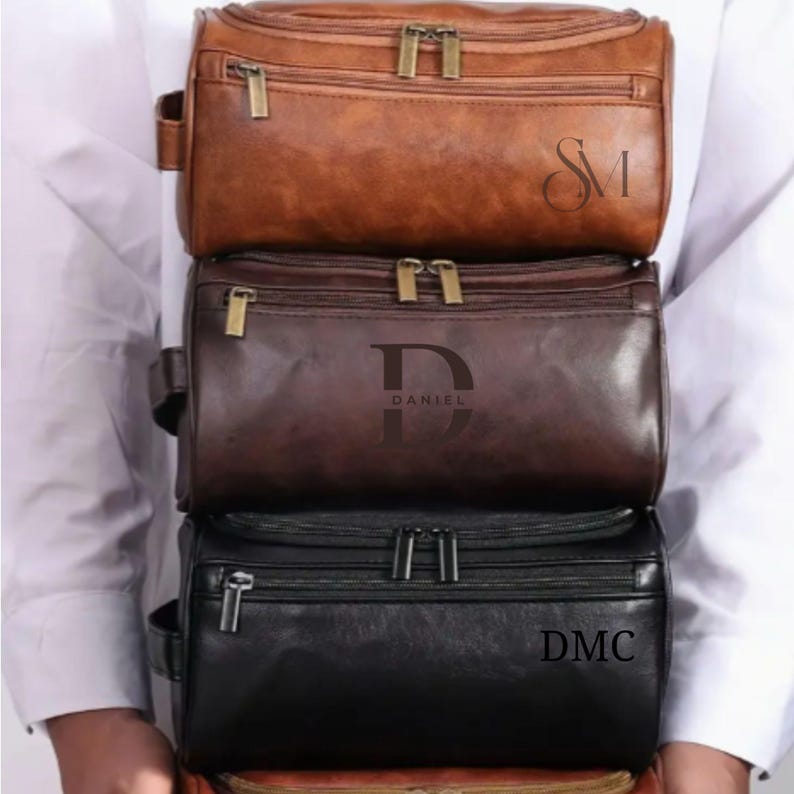 May include: Three leather wash bags stacked. The top bag is brown with the initials "SM", the middle bag is dark brown with "DANIEL", and the bottom bag is black with "DMC". Each bag has a zip closure.