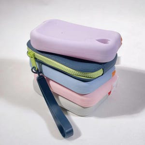 May include: A stack of five silicone pouches in various colours, including light purple, blue, pink, and grey. The pouches have a zipper closure and a strap.