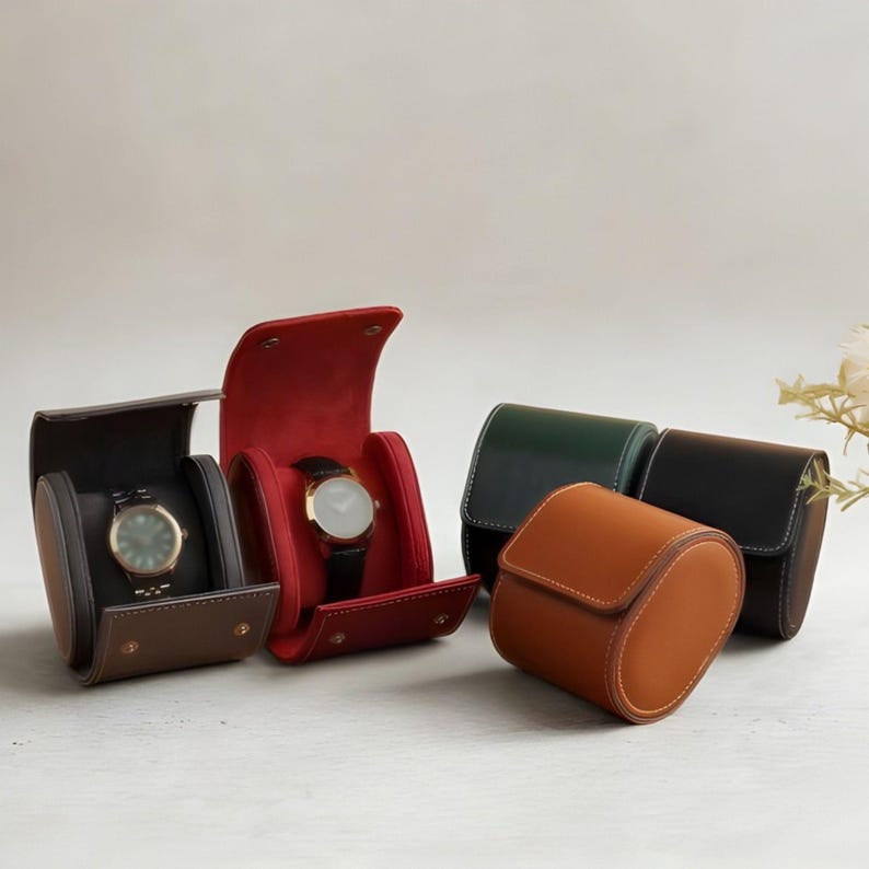 May include: A collection of cylindrical watch cases in brown, red, dark green, and black. Two cases are open, revealing interior cushions holding wristwatches with dark faces. The cases feature snap closures and visible stitching along the edges.