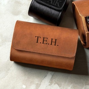 May include: A tan leather watch roll with a flap closure featuring the engraved letters T.E.H. centred on the front. The case has visible stitching along the edges and a smooth matte finish. A black leather watch roll is partially visible above.