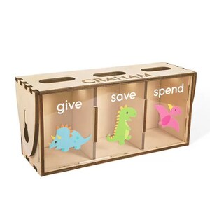 Puede incluir: Huchilla de madera con tres compartimentos etiquetados como "give", "save" y "spend". Cada compartimento tiene una ilustraci&oacute;n de dinosaurio. La huchilla est&aacute; personalizada con el nombre "GRAHAM".