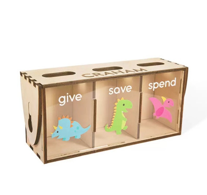 Puede incluir: Huchilla de madera con tres compartimentos etiquetados como "give", "save" y "spend". Cada compartimento tiene una ilustraci&oacute;n de dinosaurio. La huchilla est&aacute; personalizada con el nombre "GRAHAM".