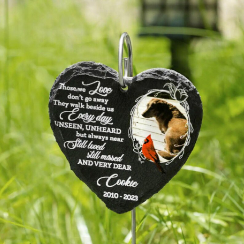 Puede incluir: Marcador de jard&iacute;n de piedra negra en forma de coraz&oacute;n con una foto de un perro y un cardenal. La piedra tiene el texto "Those we love don't go away. They walk beside us every day. Unseen, unheard but always near. Still loved, still missed and very dear. Cookie 2010-2023."