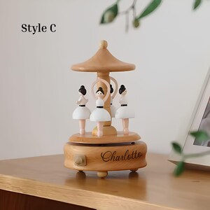 Puede incluir: Caja de m&uacute;sica de carrusel de madera con cuatro figuras de bailarinas con tut&uacute;s blancos y pelo negro. La caja de m&uacute;sica tiene un acabado marr&oacute;n claro, una parte superior decorativa y el nombre "Charlotte" grabado en la base.