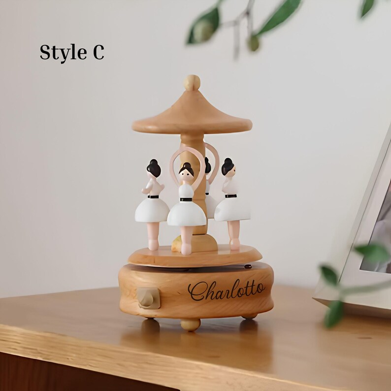 Puede incluir: Caja de m&uacute;sica de carrusel de madera con cuatro figuras de bailarinas con tut&uacute;s blancos y pelo negro. La caja de m&uacute;sica tiene un acabado marr&oacute;n claro, una parte superior decorativa y el nombre "Charlotte" grabado en la base.