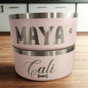May include: Two stacked light pink metal pet bowls with a silver rim. The top bowl features the name MAYA in bold silver letters with two small paw prints. The bottom bowl features the name Cali in a cursive silver script with a bone icon.