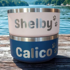 May include: Two stacked stainless steel pet bowls with a matte finish. The top bowl is light pink with the name Shelby and two paw prints engraved in silver. The bottom bowl is dark blue with the name Calico and two paw prints engraved in silver.