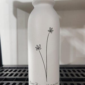 Pu&ograve; includere: Un vaso cilindrico bianco opaco con collo stretto. La parte anteriore presenta un disegno a linee nere di due semplici fiori di campo su lunghi steli. Sotto i fiori, il testo recita: Best Bonus Mom ever, Love, S.