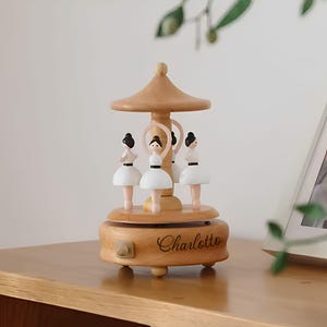 Puede incluir: Una caja de m&uacute;sica de carrusel de madera con cuatro figuras de bailarinas con tut&uacute;s blancos y cabello negro. El carrusel tiene un acabado de madera marr&oacute;n claro con el nombre "Charlotto" grabado en la base. El dise&ntilde;o incluye una parte superior en forma de hongo.