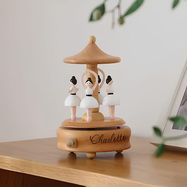 Puede incluir: Una caja de m&uacute;sica de carrusel de madera con cuatro figuras de bailarinas con tut&uacute;s blancos y cabello negro. El carrusel tiene un acabado de madera marr&oacute;n claro con el nombre "Charlotto" grabado en la base. El dise&ntilde;o incluye una parte superior en forma de hongo.