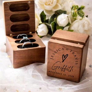 May include: A dark wood ring box with a hinged lid, featuring three oval slots containing three silver rings. The exterior of the closed box is engraved with the text JESSICA & TRAVIS, a heart symbol, the name Griffith, and NOVEMBER 16, 2026.