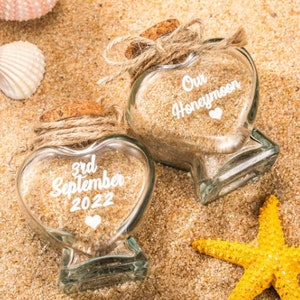 May include: Two heart-shaped glass jars filled with sand, each topped with a cork and twine. One jar features the white text Our Honeymoon with a heart icon, while the other displays 3rd September 2022 with a heart icon, resting on sand.