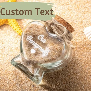 May include: A heart-shaped glass jar filled with sand, featuring a cork stopper tied with twine. The front of the jar is etched with the white text Our Honeymoon and a small heart symbol. The jar is positioned on a sandy surface with a yellow starfish.