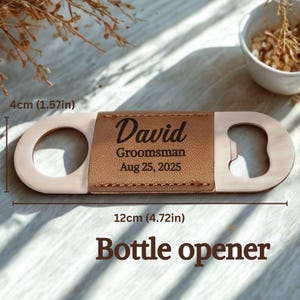 May include: A bottle opener with a tan leather center section engraved with "David, Groomsman, Aug 25, 2025." The opener is 12cm long and 4cm wide. The words "Bottle opener" are at the bottom.
