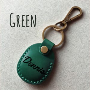Pu&ograve; includere: Portachiavi in pelle verde con il nome "Dennis" in corsivo nero. Il portachiavi ha un moschettone e un anello color oro. La parola "GREEN" &egrave; stampata in grandi lettere nere sopra il portachiavi.