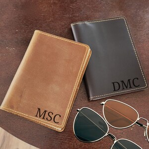 K&ouml;nnte beinhalten: Zwei Ledergeldb&ouml;rsen, eine hellbraune und eine dunkelbraune, mit personalisierten Initialen. Die hellbraune Geldb&ouml;rse hat "MSC" und die dunkelbraune Geldb&ouml;rse hat "DMC". Eine Sonnenbrille mit silbernem Gestell und braunen Gl&auml;sern ist ebenfalls im Bild.