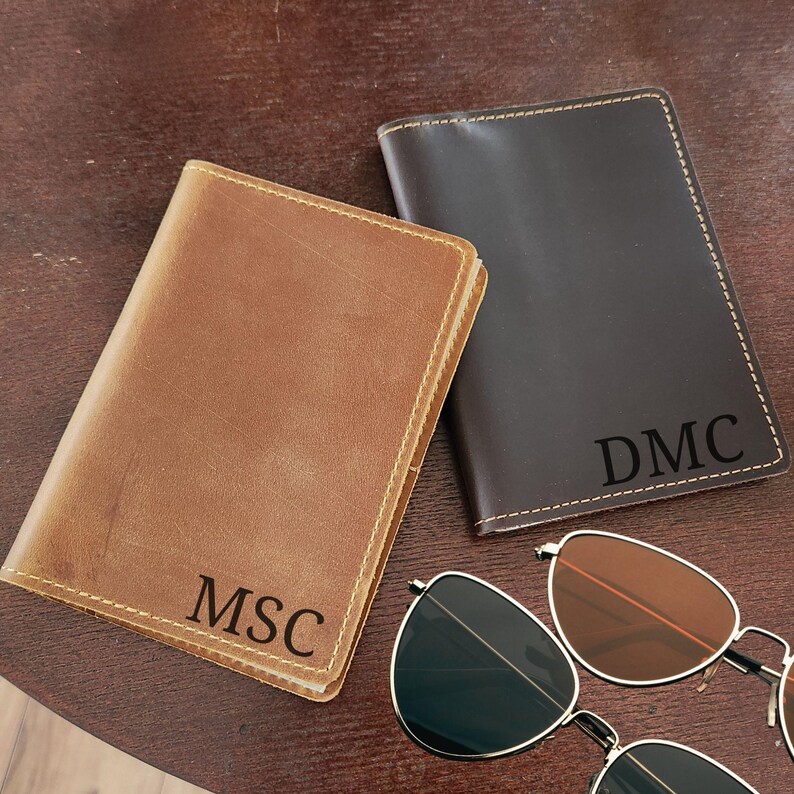 K&ouml;nnte beinhalten: Zwei Ledergeldb&ouml;rsen, eine hellbraune und eine dunkelbraune, mit personalisierten Initialen. Die hellbraune Geldb&ouml;rse hat "MSC" und die dunkelbraune Geldb&ouml;rse hat "DMC". Eine Sonnenbrille mit silbernem Gestell und braunen Gl&auml;sern ist ebenfalls im Bild.