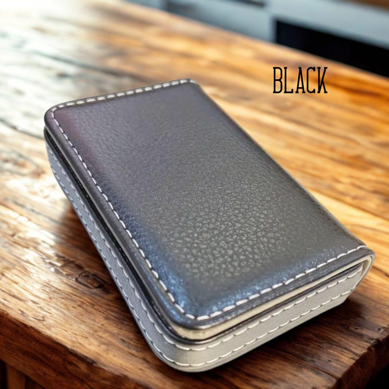 May include: A black, rectangular case with white stitching around the edges. The word "BLACK" is printed in the upper right corner. The case is sitting on a wooden surface.