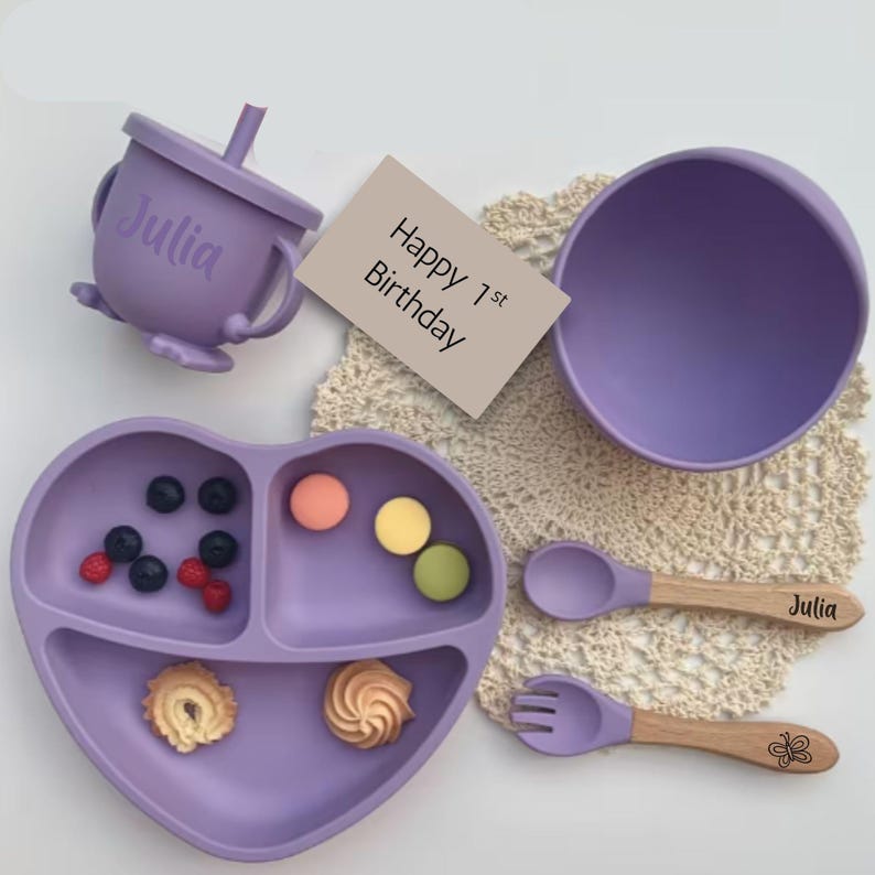 May include: A lavender baby feeding set with a heart-shaped divided plate, bowl, cup, spoon, and fork. The spoon and fork have wooden handles. The cup and spoon are personalized with the name "Julia". The plate contains berries and cookies. A card reads "Happy 1st Birthday".