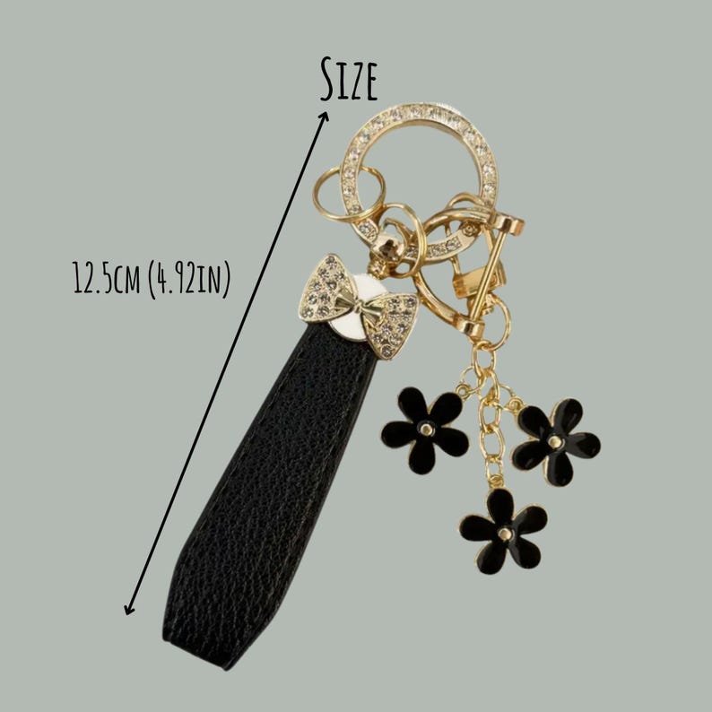 Op de afbeelding: Een goudkleurige sleutelhanger met een zwarte leren band, een met strass bezette strik en drie zwarte bloembedels. De sleutelhanger is 12,5 cm lang. Het woord "SIZE" staat boven de sleutelhanger.