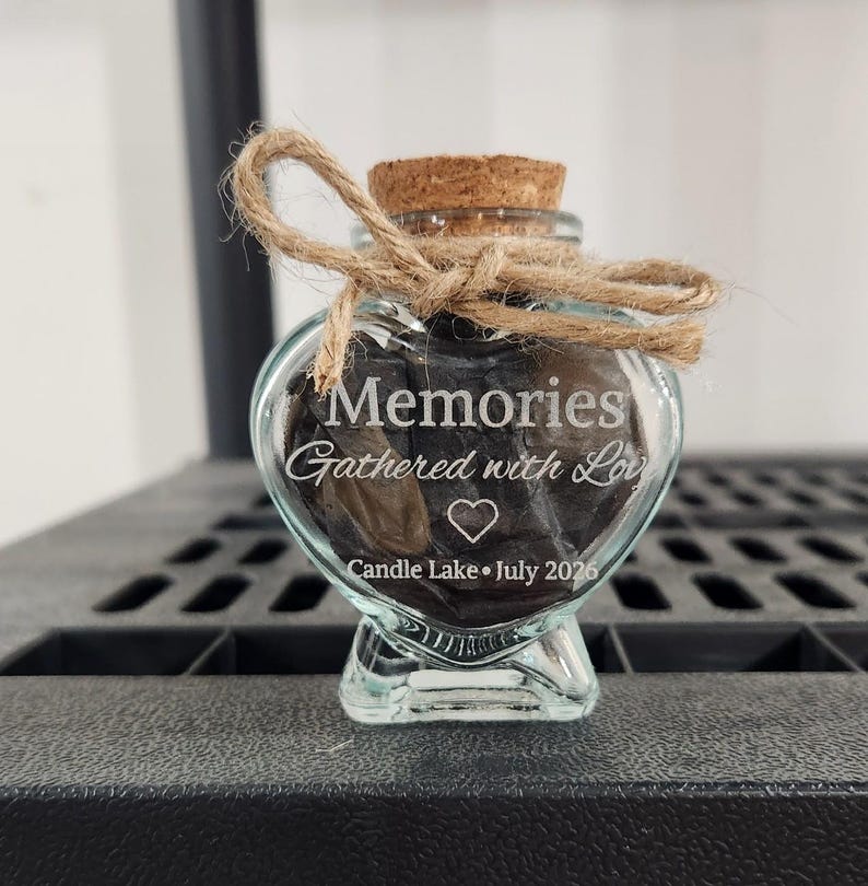 May include: A heart-shaped glass bottle with a cork stopper and a tied jute twine bow. The glass is etched with the text Memories Gathered with Love, a heart icon, and the text Candle Lake July 2026. The bottle contains dark crumpled paper.