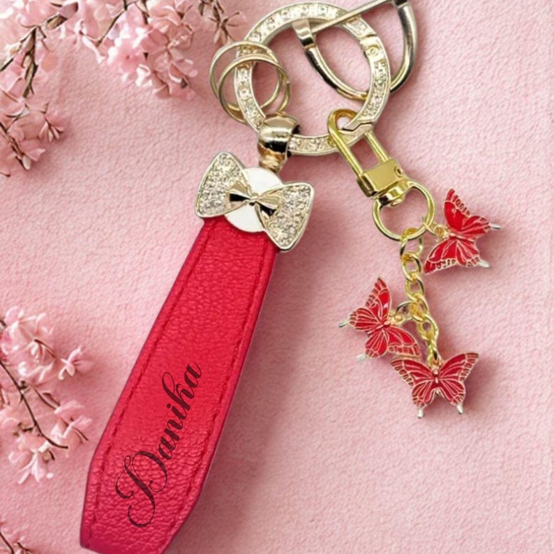 Op de afbeelding: Een rode leren sleutelhanger met de naam "Danika" in cursief schrift. De sleutelhanger heeft een strikvormige bedel met strass accenten en een goudkleurige sluiting. Drie rode vlinderbedels hangen aan de sleutelhanger.