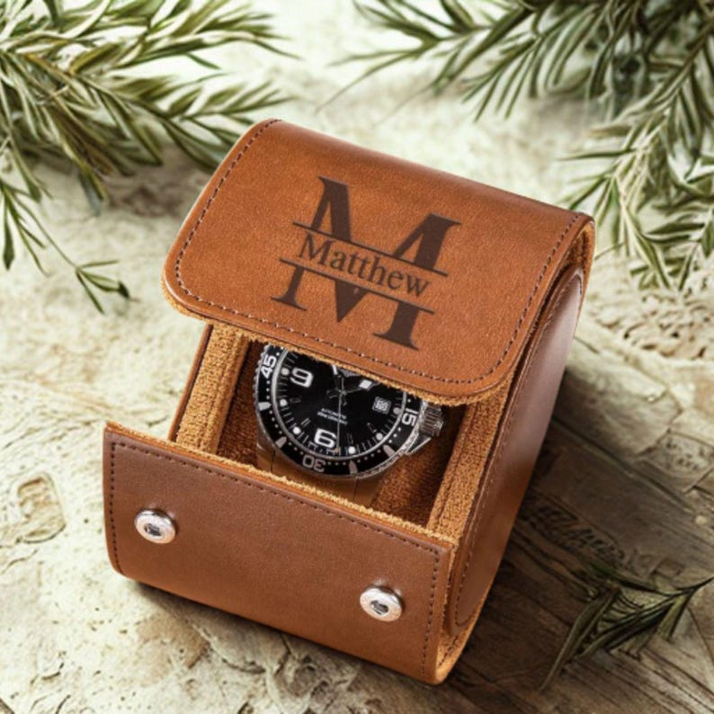 May include: A tan leather watch roll with a hinged lid featuring the engraved letter M and the name Matthew. The interior is lined with a soft tan material and holds a silver-toned wristwatch with a black dial and a rotating bezel.