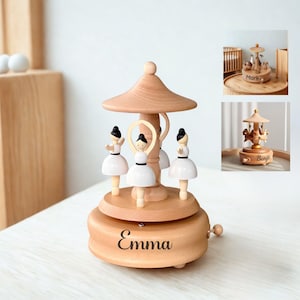 Puede incluir: Una caja de m&uacute;sica de carrusel de madera con tres figuras de bailarina con tut&uacute;s blancos y pelo negro. El carrusel tiene un acabado de madera marr&oacute;n claro con una parte superior decorativa en forma de paraguas. El nombre "Emma" est&aacute; impreso en la base.