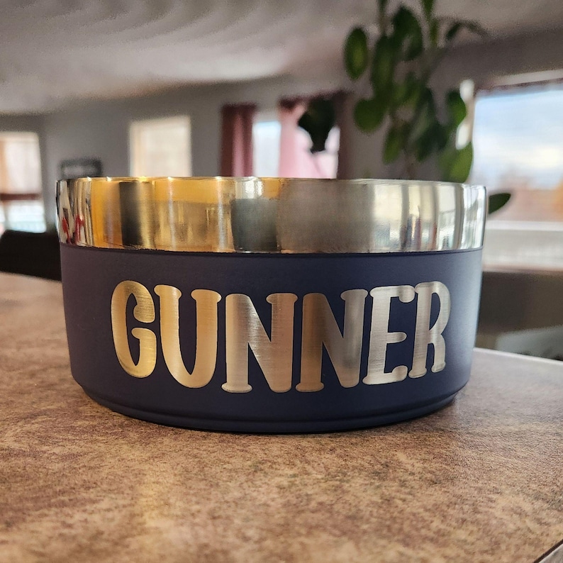 May include: A round, navy blue pet bowl with a metallic silver rim. The name GUNNER is engraved in large, bold, capital letters across the front of the bowl, revealing a metallic silver finish beneath the matte navy blue exterior coating.