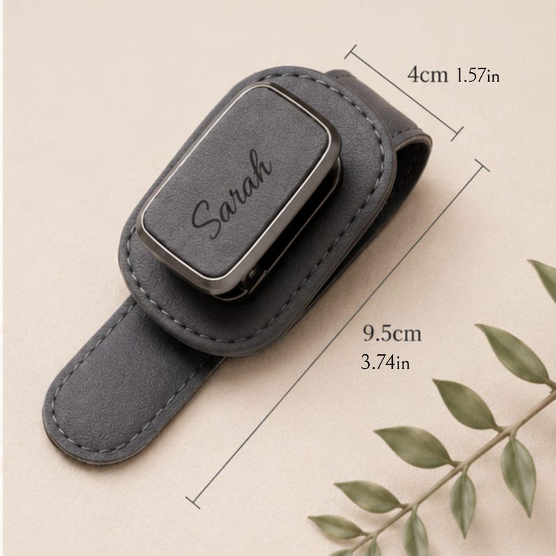 K&ouml;nnte beinhalten: Ein dunkelgrauer magnetischer Lederclip mit dem eingravierten Namen Sarah auf der vorderen rechteckigen Oberfl&auml;che. Der Clip ist 4 cm breit und 9,5 cm lang und weist sichtbare N&auml;hte entlang der Kanten der Lederbasis auf.