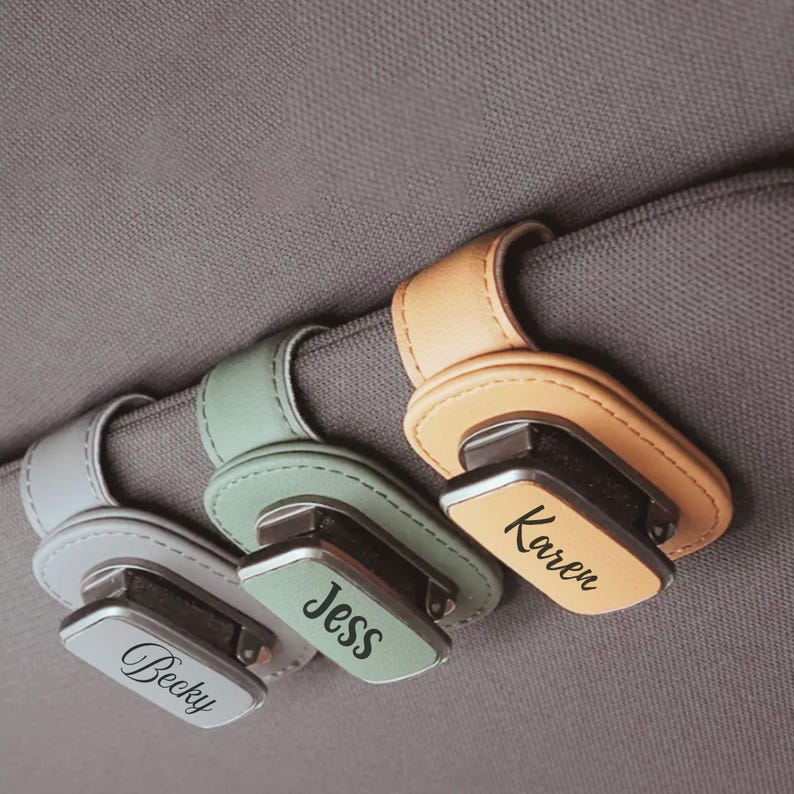 K&ouml;nnte beinhalten: Drei Sonnenbrillenhalter f&uuml;r die Sonnenblende im Auto aus hellgrauem, salbeigr&uuml;nem und beigefarbenem Kunstleder. Jeder Halter hat einen Metallclip mit einem in schwarzer Schreibschrift eingravierten Namen: Becky, Jess und Karen auf Stoff.