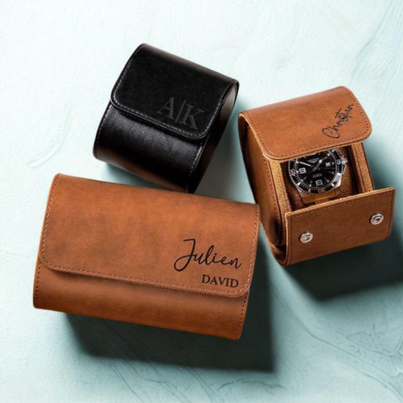 May include: Three leather watch cases in black and brown. One black case has "AK" engraved on the flap. A brown case displays a watch. Another brown case is engraved with "Julien DAVID". The cases are on a light blue surface.