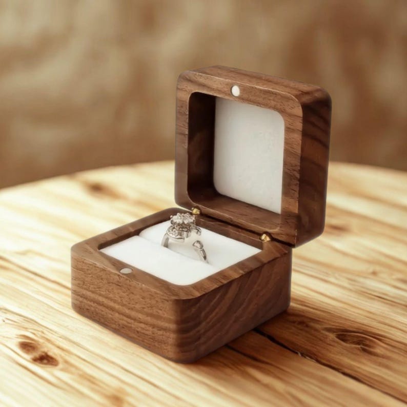 May include: Open wooden ring box with a white interior, showcasing a silver ring with a large, ornate gemstone. The box is made of dark wood and sits on a light wooden surface. The ring is the focal point.