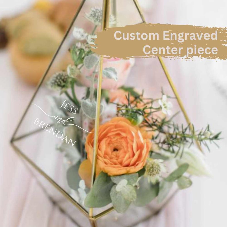 K&ouml;nnte beinhalten: Ein geometrisches Glasterrarium mit goldenem Metallrahmen, gef&uuml;llt mit orangefarbenen und rosafarbenen Blumen sowie Gr&uuml;npflanzen. Das Glas ist mit dem Text JESS and BRENDAN graviert. Ein beigefarbenes Overlay zeigt Custom Engraved Center piece.