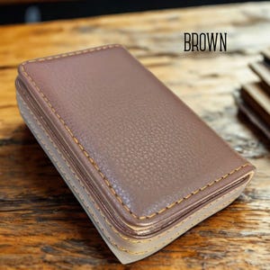May include: A brown leather business card holder with gold stitching. The word "BROWN" is printed in black at the top. The holder has a textured surface and a rectangular shape.