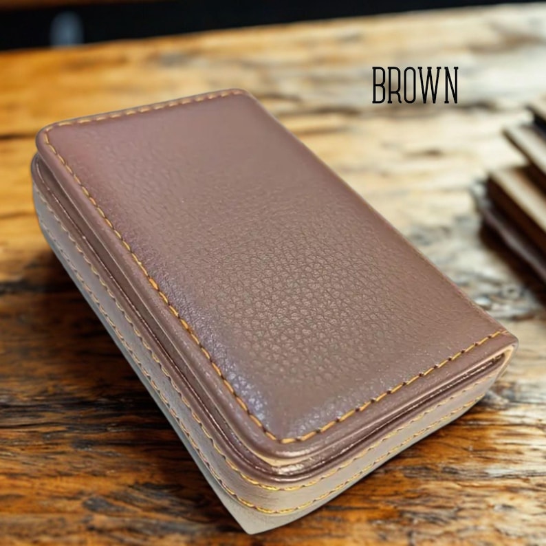 May include: A brown leather business card holder with gold stitching. The word "BROWN" is printed in black at the top. The holder has a textured surface and a rectangular shape.