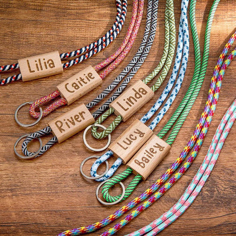 Pode incluir: Um conjunto de chaveiros tran&ccedil;ados coloridos com etiquetas de madeira gravadas com nomes como "Lilia", "Caitie", "River", "Linda", "Lucy" e "Bailey".